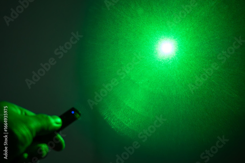 A person's hand holding a remote with a glowing blue light and a green laser beam pointing upwards in a hazy atmosphere.