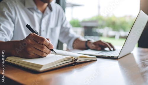 A person diligently working at a wooden desk, taking notes in a notebook while using a laptop computer.