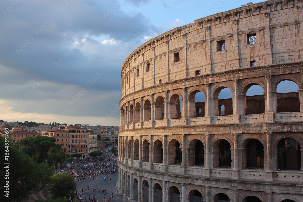 Fototapeta premium Colosseum in Rome stands majestically against epic sky, surrounded by ancient ruins and bustling crowds. historic architecture is beautifully illuminated by setting sun