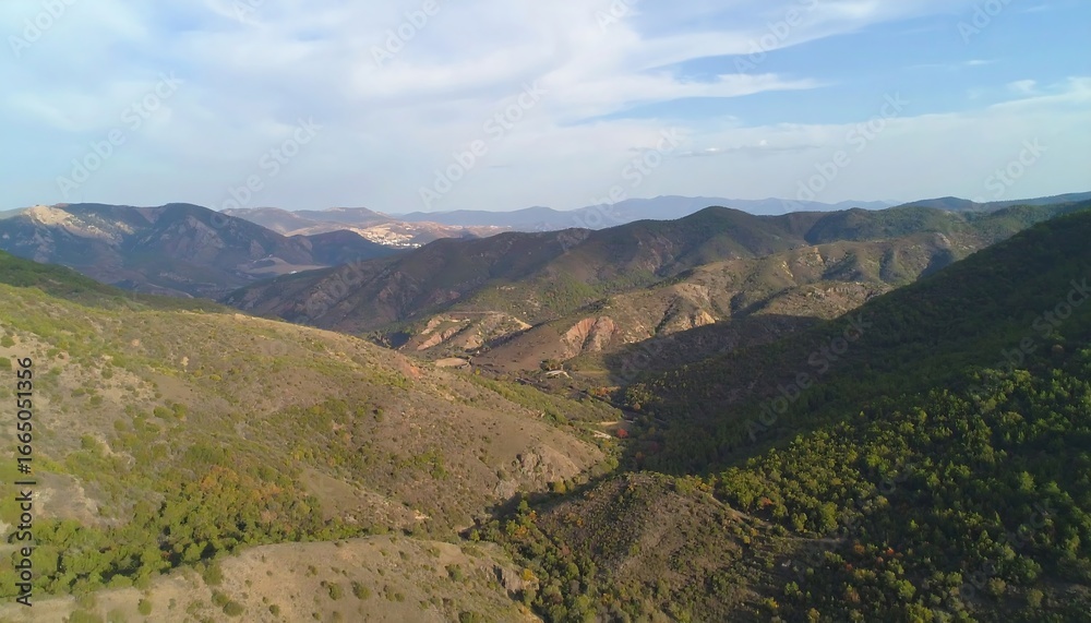 Fototapeta premium A high-angle view reveals a valley nestled between rolling hills, showcasing a mix of light brown and green vegetation, and a pale blue sky.