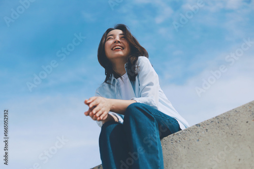 Cheerful young Asian woman sitting  on the roof under blue sky background.