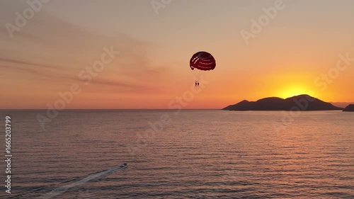 A breathtaking 4K aerial shot of two people parasailing in Oludeniz Fethiye their parachute with a Turkish flag silhouetted against a spectacular golden sunset over the calm sea