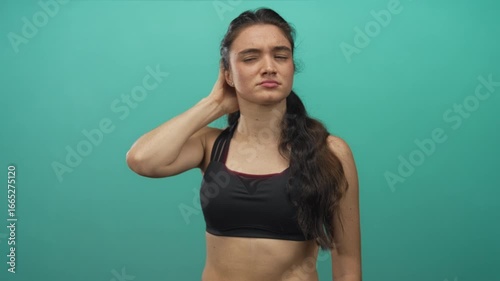 Hispanic woman in black sports bra touching nape and neck with closed eyes against a turquoise studio wall; discomfort.