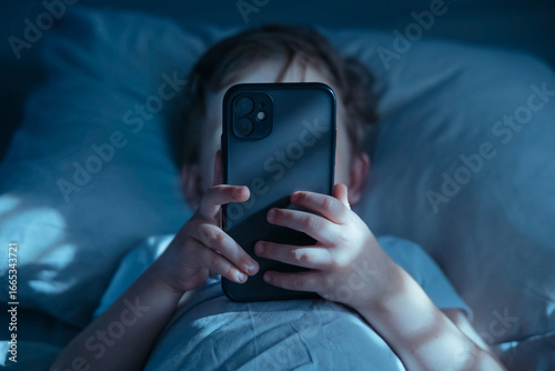 Close-up of unrecognisable little child lying in bed at night, holding mobile phone in front of his face. Gadget addiction and excessive late-night screen time. Smartphone overuse 