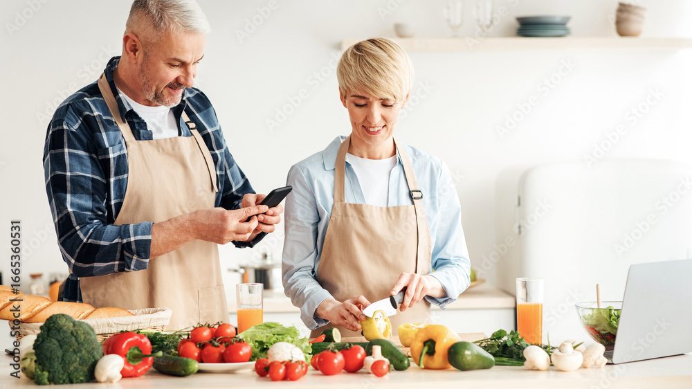 © Prostock-studio - Cook delicious meal together, healthy eating and vegetarian dish. Happy senior woman in apron prepare food, man makes photo on smartphone in kitchen interior with bright vegetables and juice on table