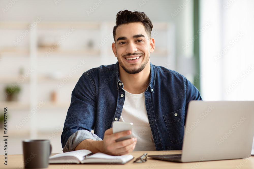 Obraz premium Smiling arab guy freelancer using smartphone while sitting at workdesk at home, working on laptop, copy space. Cheerful handsome middle-eastern man holding mobile phone, office interior
