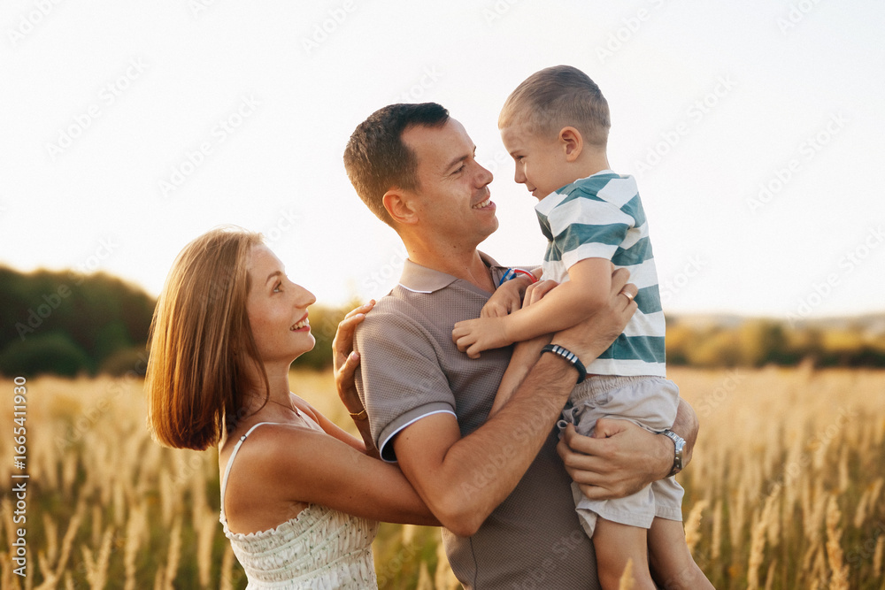 Fototapeta premium A joyful family spends time outdoors in a golden field during late afternoon. The father holds his smiling son, while the mother looks at them with affection.