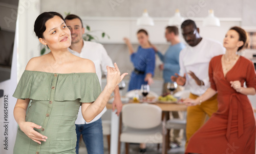 Smiling carefree Asian woman in olive ruffled dress cheerfully dancing surrounded by friends at laid-back home dance party..