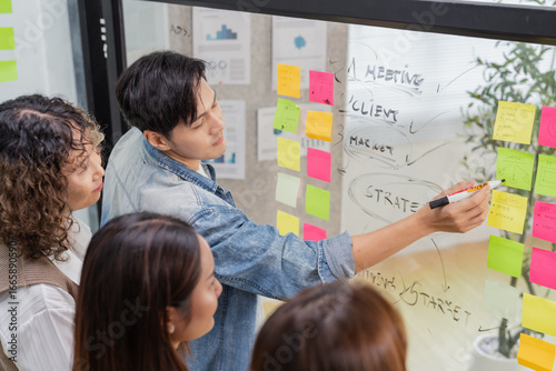 Asian business creative marketing team brainstorming startup strategy using glassboard and sticky notes. Collaborative ideation session demonstrates design thinking methodology disruptive innovation