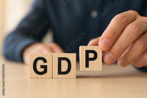 A person's hand carefully places a wooden block displaying the letter 'P' next to blocks spelling out 'GDP' on a light beige wooden table.