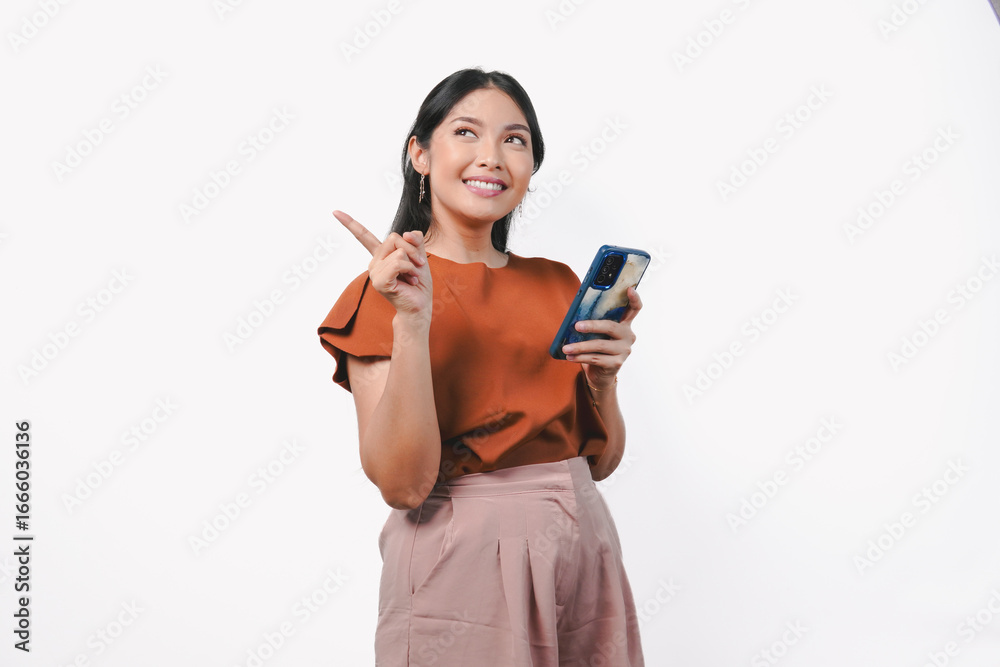 Cheerful young Asian woman holding smartphone while pointing to the copy space behind her. Cheerful young Asian woman holding smartphone while pointing to the copy space behind her.