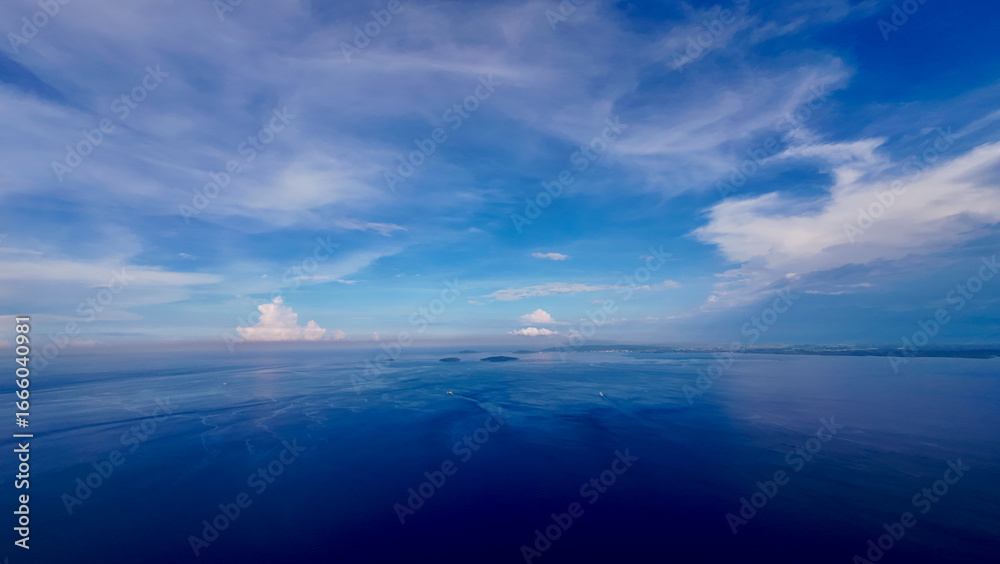 Fototapeta premium Aerial view of a flat calm sea surface and white clouds. Small clouds above the ocean surface. View from above of a calm ocean surface, small tropical islands and ships moving on the sea.