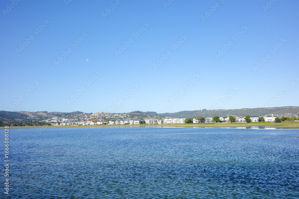 Fototapeta premium A calm lagoon with gentle rippling water reflecting the bright blue sky, with a charming coastal town and hills as backdrop. The peaceful view with scenic view of travel and lifestyle