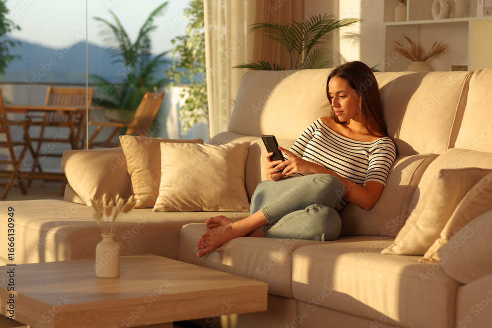 Obraz premium Relaxed woman at sunset at home using cell phone on a couch