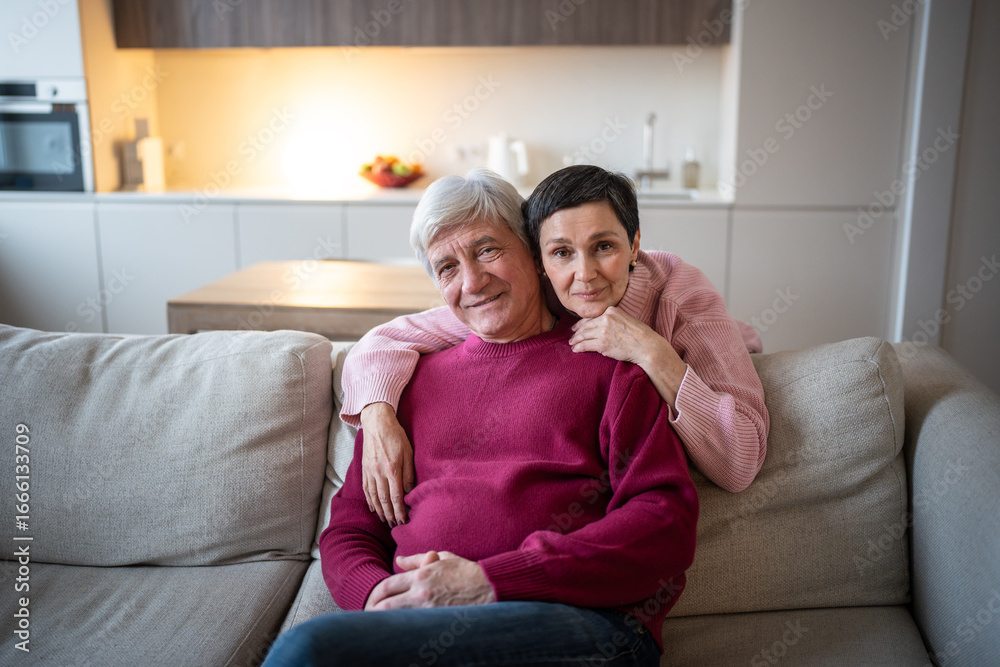 Obraz premium Smiling senior married couple hugging on sofa at home looking at camera. Happy family love and care, trust bonding in relationship, loving happy old husband and wife embracing behind in apartment