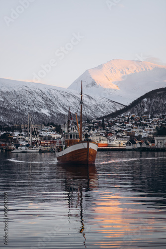 A classic wooden boat navigates the calm waters of the Tromsø fjord, with the city's buildings and majestic, snow-covered mountains illuminated by the setting sun in the background.