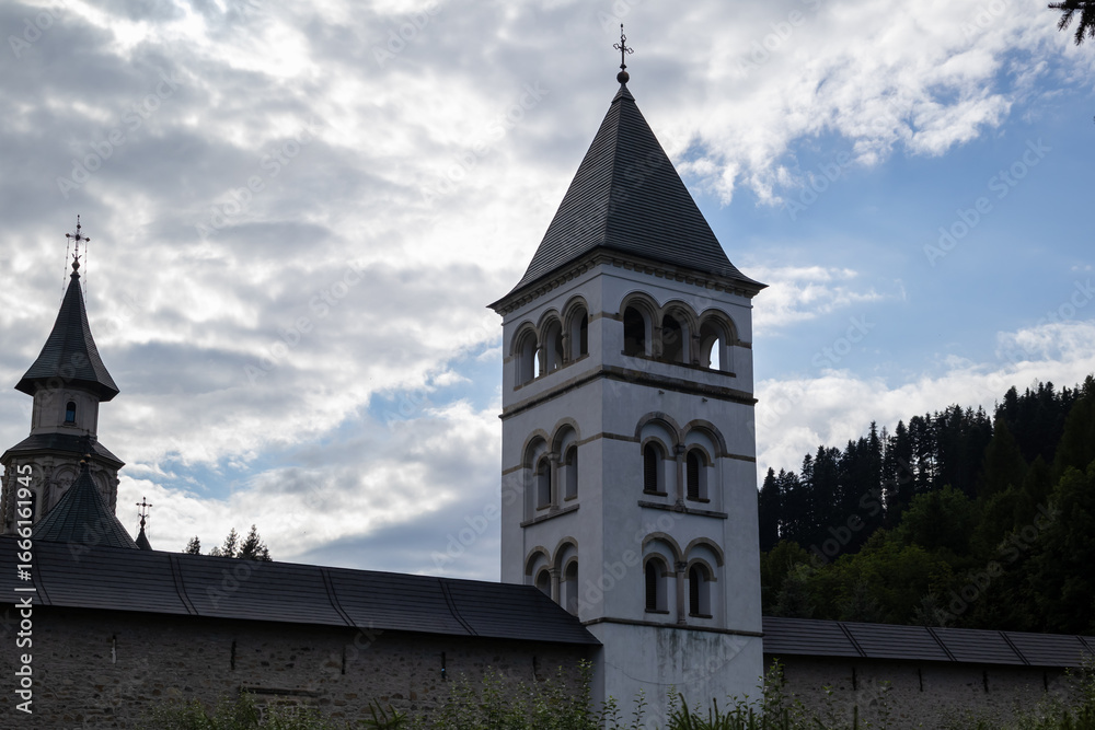 Fototapeta premium a low-angle shot of the iconic Putna Monastery in Romania, featuring the majestic bell towers and the stone fortified wall under a dramatic sky with clouds.