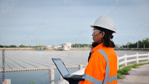 A woman in a hard hat and reflective jacket assesses a water treatment plant, using her laptop for data analysis. The facility features numerous large tanks under an overcast sky