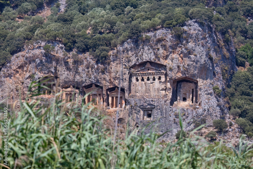 Carved Lycian tombs sit high in a rocky cliff face, surrounded by green vegetation. The tombs have columned facades and dark rectangular openings - Caunos Tombs of the Kings - Dalyan - Mugla - Turkey Carved Lycian tombs sit high in a rocky cliff face, surrounded by green vegetation. The tombs have columned facades and dark rectangular openings - Caunos Tombs of the Kings - Dalyan - Mugla - Turkey