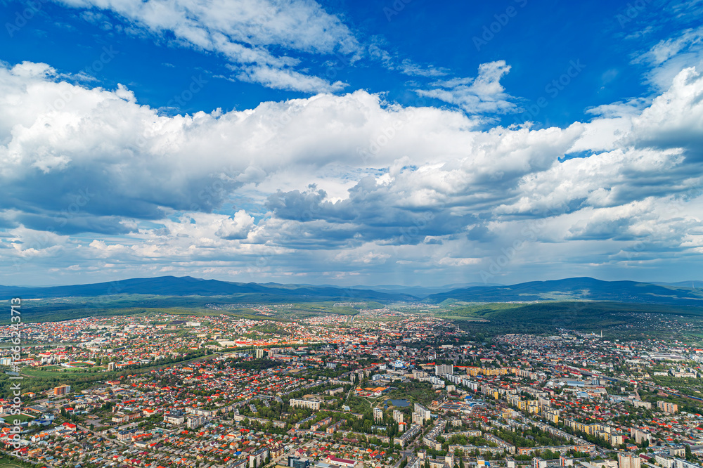 Fototapeta premium An aerial panoramic view of a city surrounded by mountains and greenery under a dramatic cloudy sky.