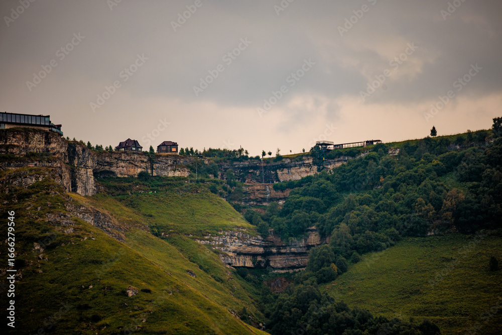 Naklejka premium A scenic view of a rocky cliff in the Caucasus region, featuring lush greenery and a small waterfall. A viewing platform is visible on the cliff edge.