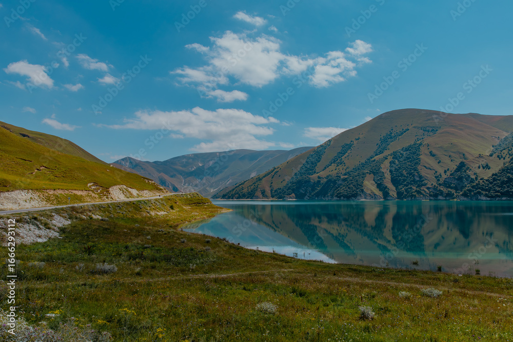 Naklejka premium A serene landscape in the Caucasus mountains featuring a calm lake reflecting the surrounding hills and blue sky. Lush green grass lines the shore.