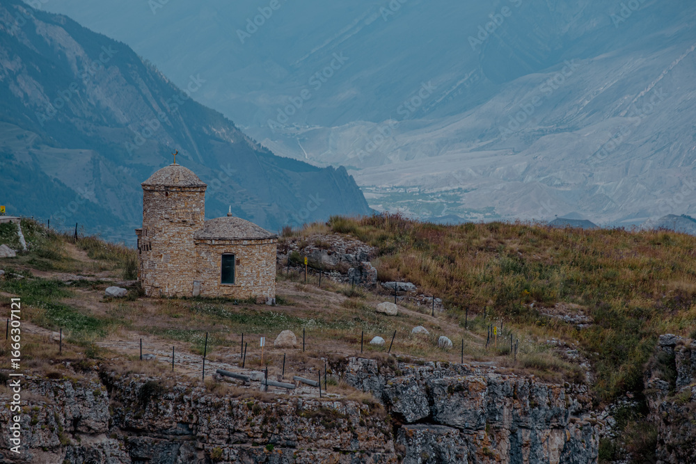 Fototapeta premium A historic stone building sits on a rocky hillside in the Caucasus Mountains. The landscape features rugged terrain and distant peaks under a cloudy sky.