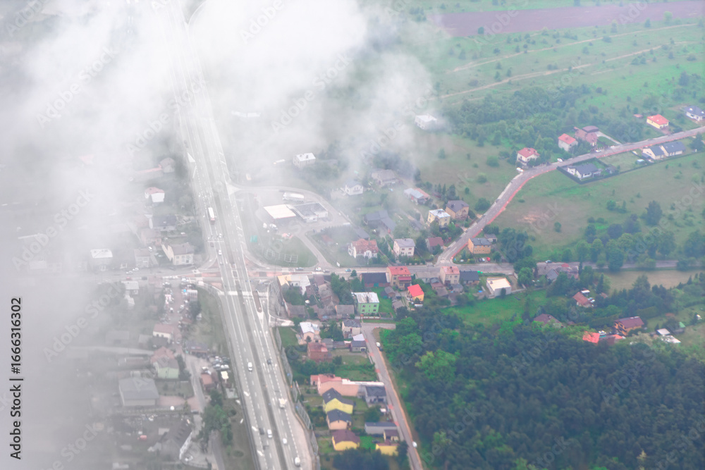 Naklejka premium Aerial view of small town partially covered by drifting clouds reveals roads, houses and green fields. Scenic perspective captures rural charm and contrast between urban layout and natural landscape