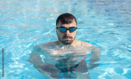 Man with swim goggles resting with elbows on pool edge, water dripping from face. Male emerging from fresh blue water in summer sunlight. Relaxed vacation moment, healthy lifestyle and leisure.