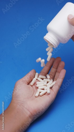 White capsules or pills being poured from a plastic bottle into a hand against a blue background. Concept of medicine, healthcare, supplements, and pharmaceutical treatment.