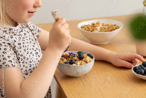The girl has granola with yogurt and berries for breakfast on the table. 