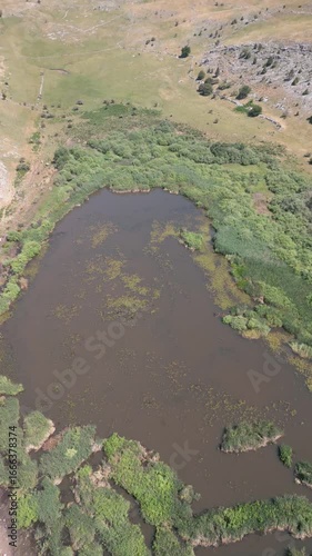 Drone aerial footage of a small mountain lake surrounded by rocky hills and green vegetation, representing untouched nature and scenic landscape.