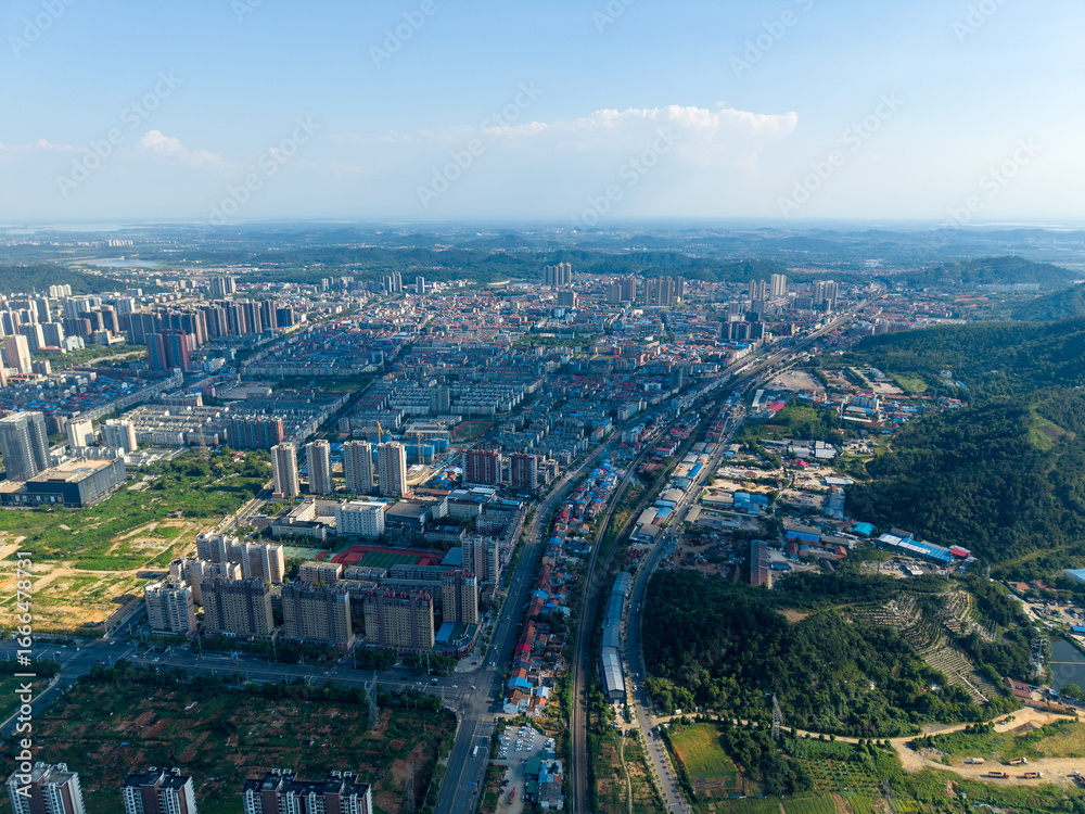 Obraz premium An expansive aerial view displays the sprawling urban panorama of Jiangxia, Wuhan. Numerous structures rise beside green hills, extending into the natural surroundings under a bright clear sky.