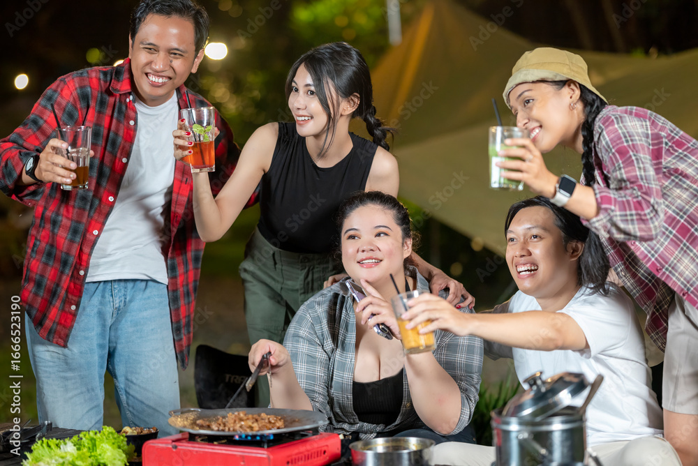 Fototapeta premium Indonesian southeast asian people relaxing together outdoors and enjoying refreshing drinks while cooking sliced beef on a grill pan. The scene of the lifestyle of enjoying food in a natural setting