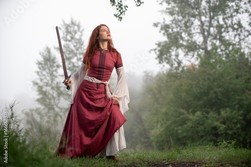 Schilderij op canvas Redhead warrior gazes skyward, sword in hand, amidst misty woodland scenery