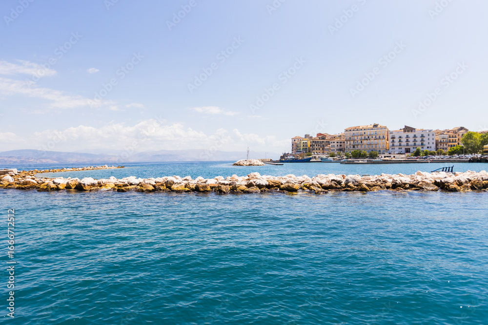 Fototapeta premium Picturesque Corfu Town harbor featuring rows of white beach umbrellas along stone breakwater with colorful Venetian buildings and crystal clear turquoise Ionian Sea waters.