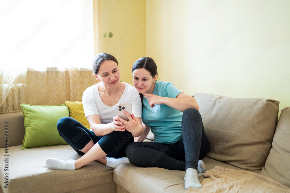 Fototapeta premium Two female friends laughing on the couch while looking at a smartphone, discussing something funny, concept of female friendship and emotional support