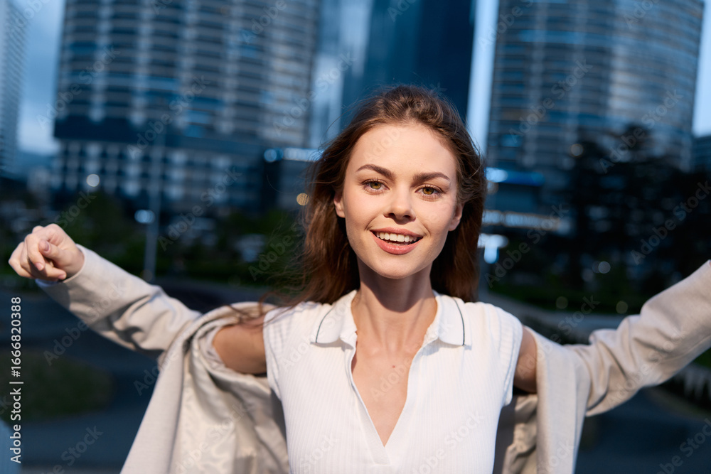 Young woman in white shirt smiling outdoors with open coat and city buildings in background at dusk, positive energy, casual style, urban fashion Young woman in white shirt smiling outdoors with open coat and city buildings in background at dusk, positive energy, casual style, urban fashion