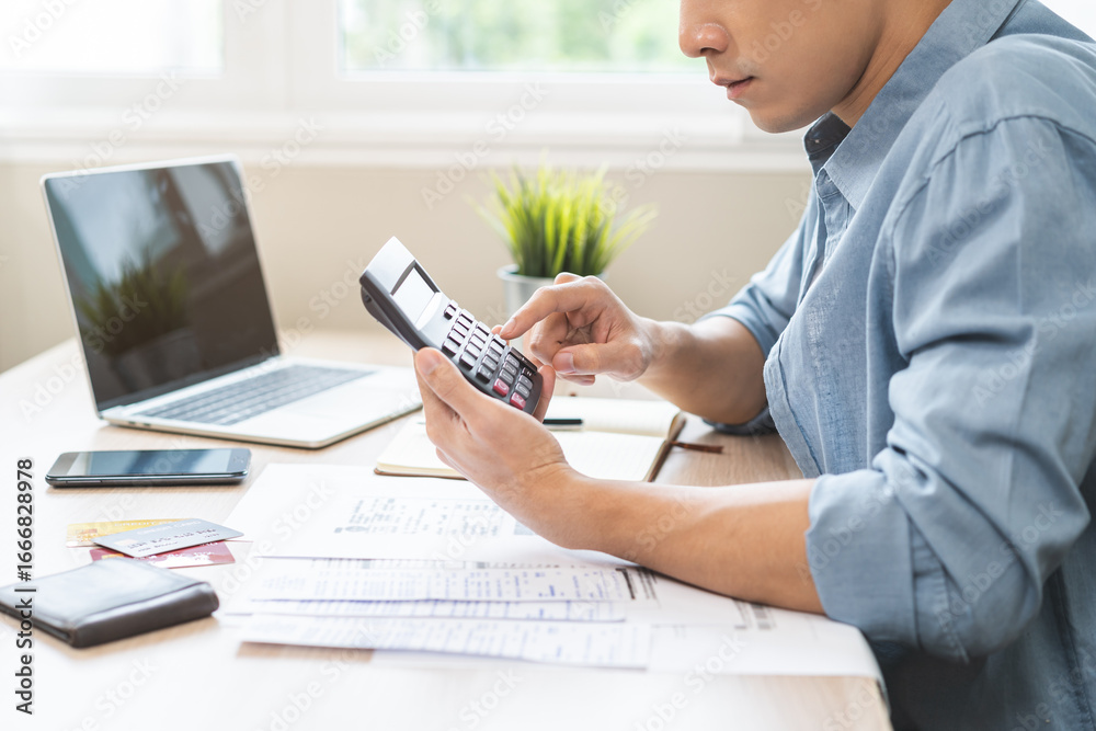 Fototapeta premium Saving money wealth concept, asian young business man, male using calculator for calculate saving cost, cash finance planning to spend enough money on his income for save money, payment tax, investing
