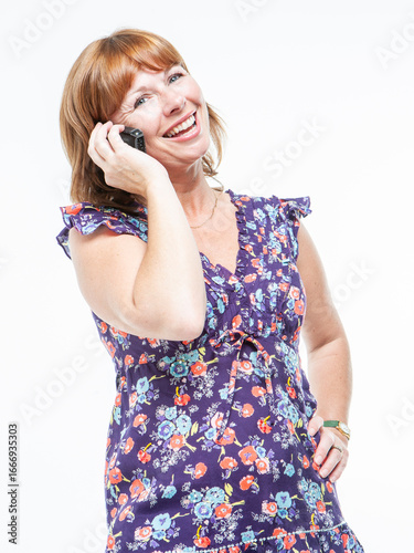 Phone Conversation. A mature woman talking on her mobile phone with a wide smile on her face. She is looking to the camera with a happy expression and wearing a purple floral dress.