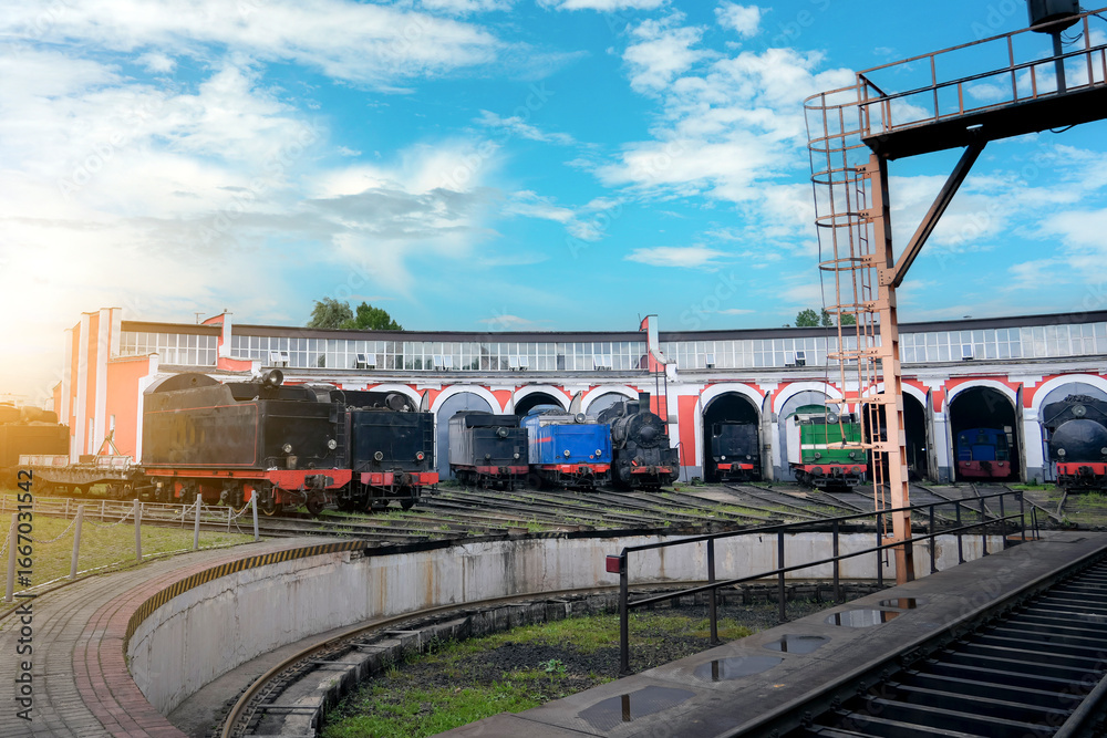 Naklejka premium Many old steam locomotives are parked inside a vintage red and white roundhouse in Eastern Europe. Tracks and maintenance equipment surround the trains