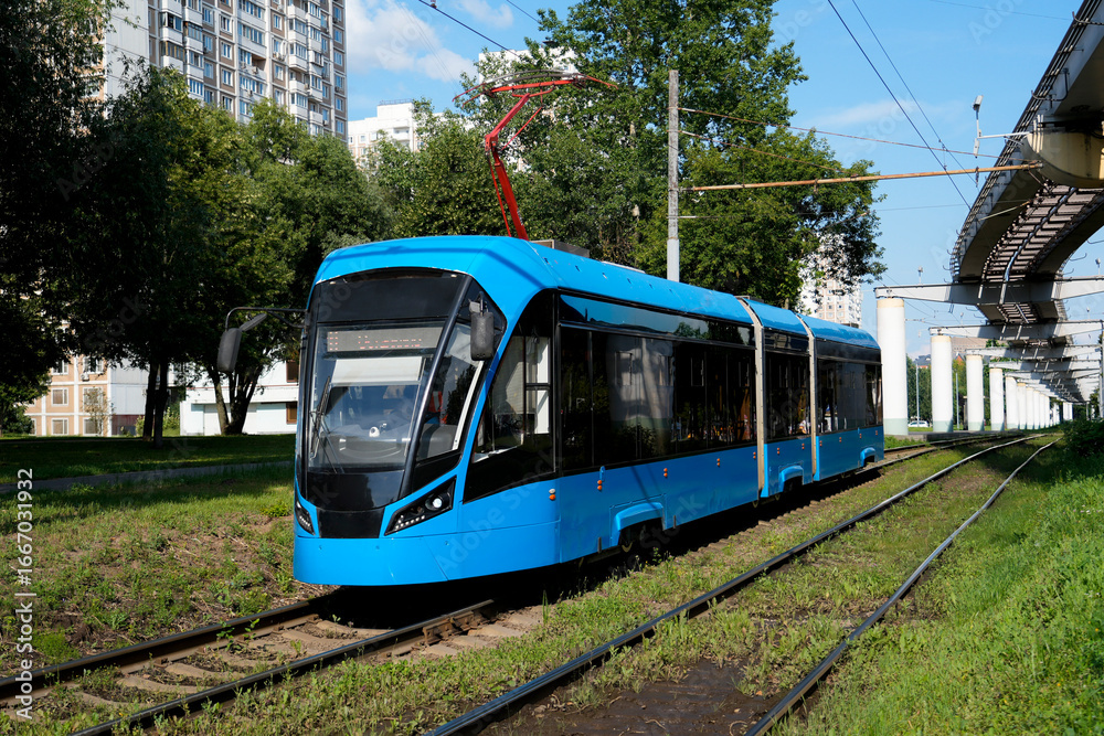 Naklejka premium A bright blue tram moves along its tracks in an urban setting on a sunny day. Apartment buildings and an elevated structure are visible in the background
