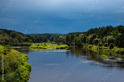 A calm river winding through lush green forests and meadows under a dramatic blue stormy sky.