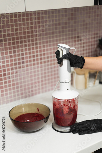 Close-up of a woman's gloved hands cooking berry jam made from fresh raspberries in the kitchen with a blender