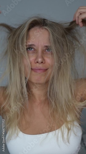 Vertical video of a young upset blonde woman looking at her tangled unruly dry bleached hair holding in her hands on a grey background. Electrifying porous hair