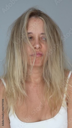 Vertical video of a young upset blonde woman touching her tangled unruly dry bleached hair holding in her hand on a light grey background. Hair care