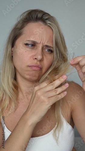 Vertical video of a young caucasian blonde woman looking at the bleached brittle split ends of her hair on a grey background. Hair care