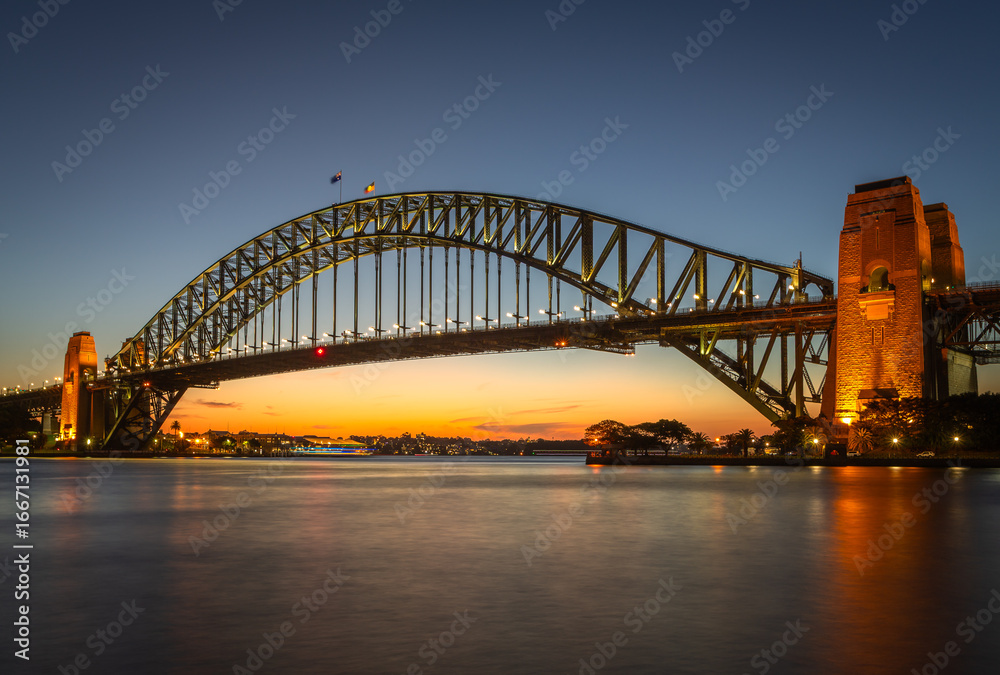 Fototapeta premium Iconic Sydney Harbour Bridge at twilight, beautifully lit with reflections on the water, under a golden sunset sky in Australia’s most famous harbor