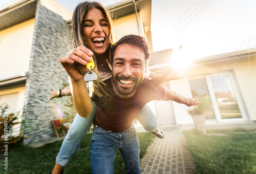 Happy young couple holding home keys after buying real estate - Husband and wife standing outside in front of their new house	