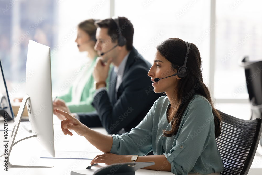 Fototapeta premium Focused young woman wear headset sit in modern office, pointing at pc screen, engaged in conversation, providing support or assistance to client as customer service representative or technical advisor